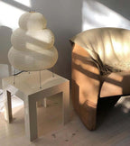 This Clérachic tiered paper lamp stands on a white side table next to a tan leather armchair, lit by afternoon sun.