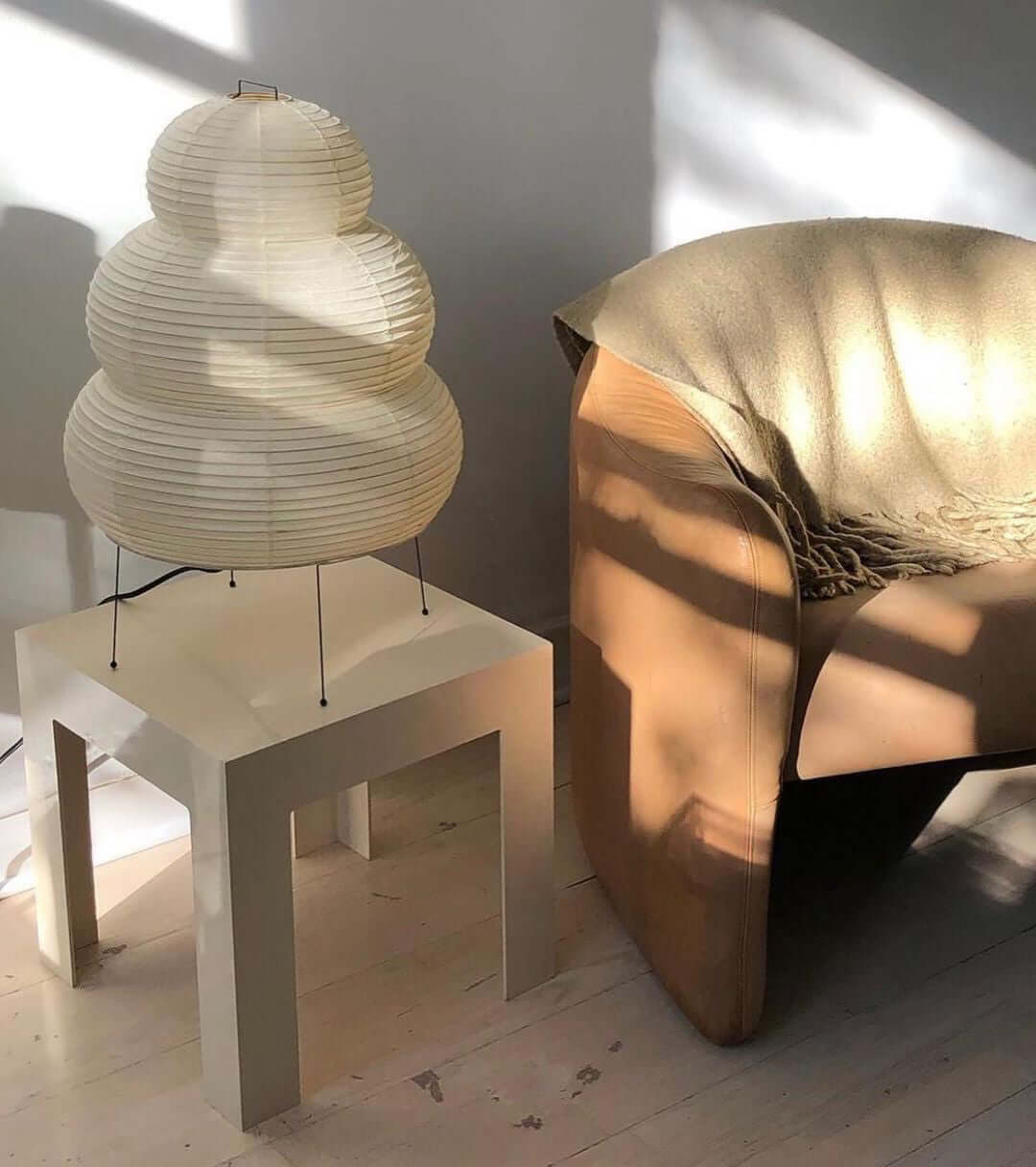This Clérachic tiered paper lamp stands on a white side table next to a tan leather armchair, lit by afternoon sun.