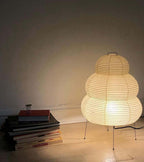 A Clérachic tiered paper lamp lit on the floor beside a stack of art books, its warm glow spreading over the wall.