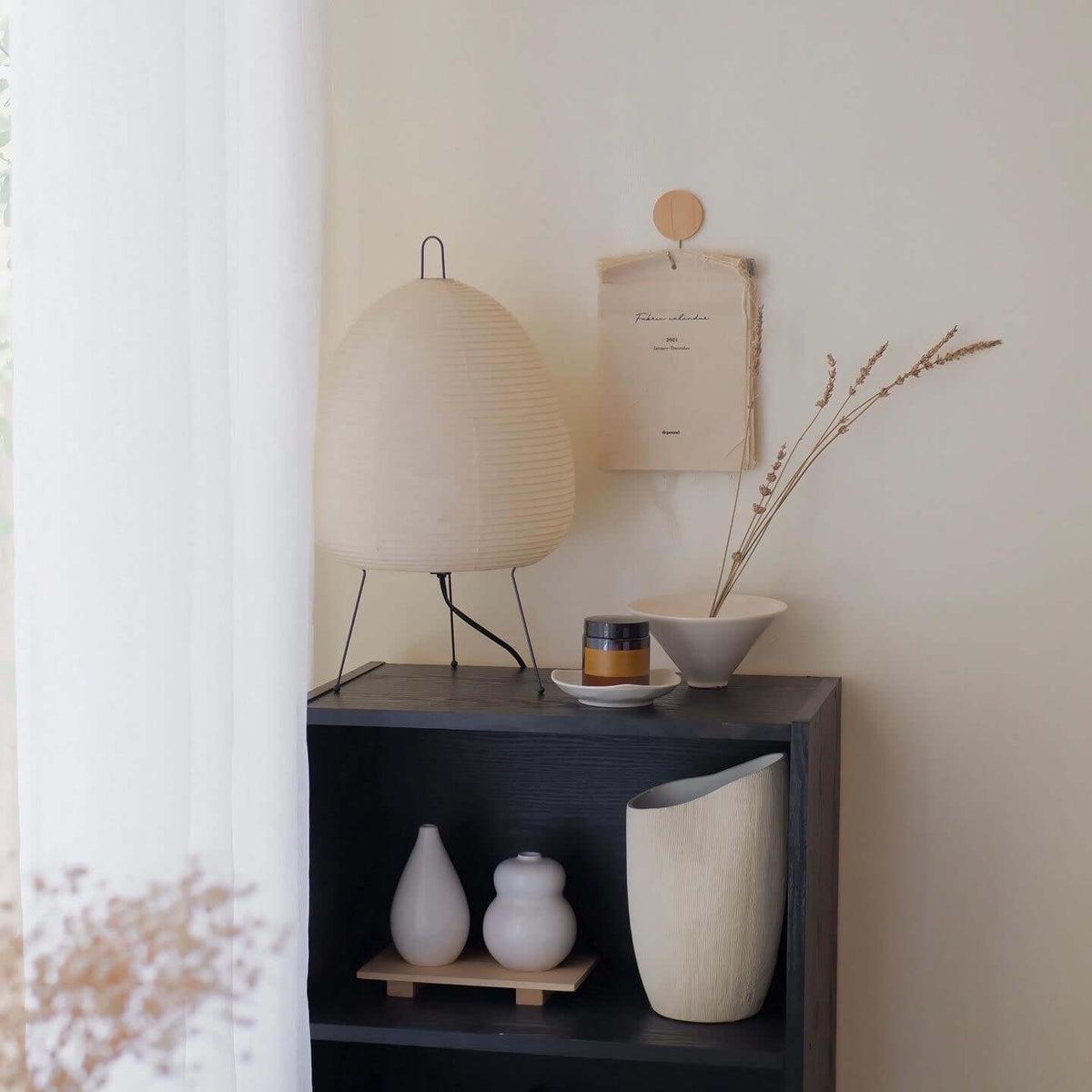 Egg-shaped Clérachic paper lamp, unlit, on a black shelf unit with ceramic vases and dried grasses nearby.