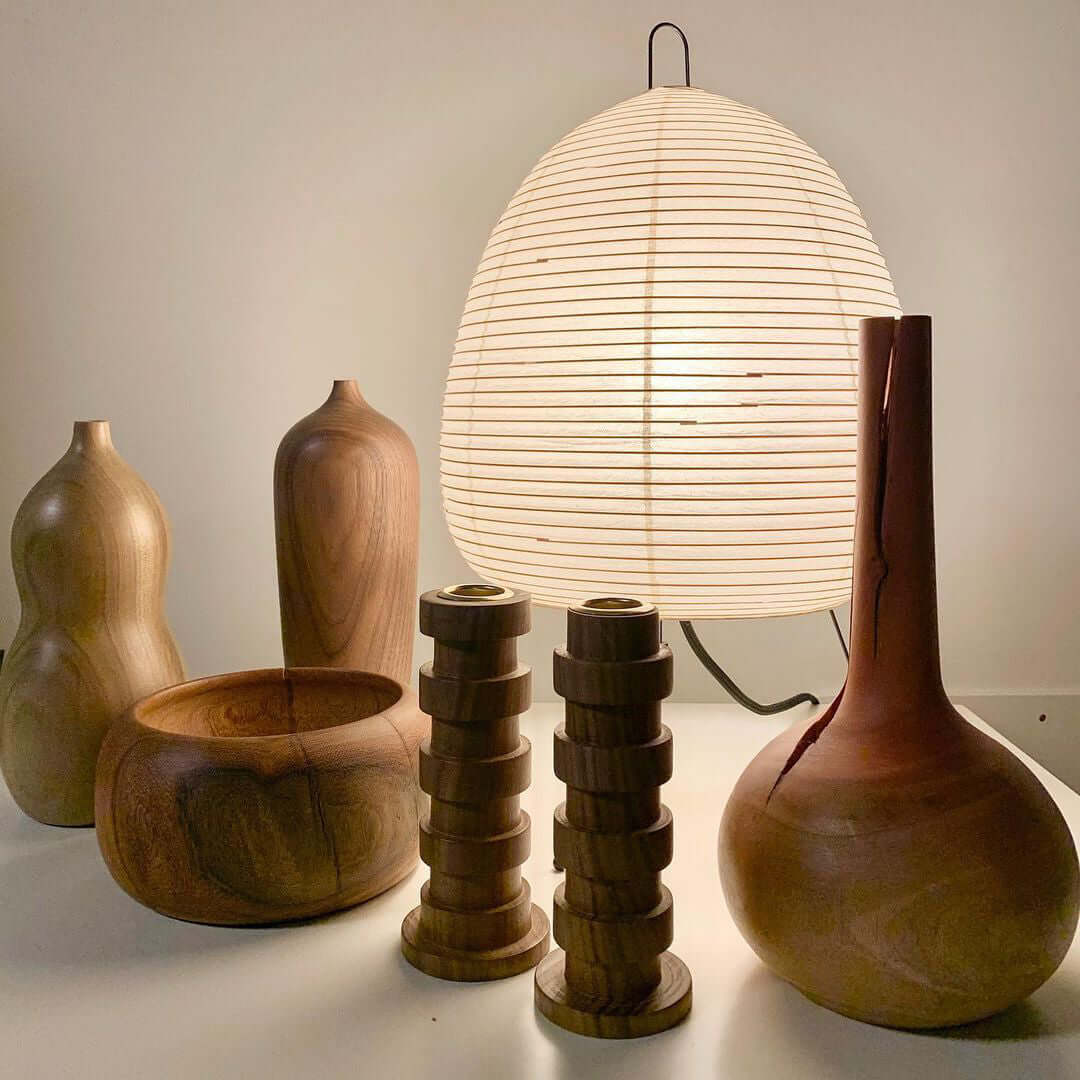 A Clérachic paper lamp glowing on a white table among turned wooden vases, bowls and candle holders.