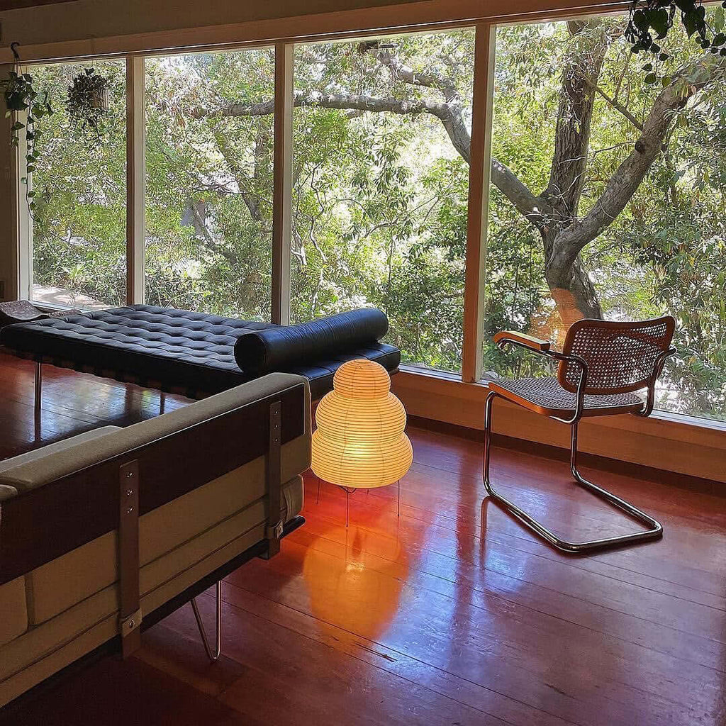 Small Clérachic tiered paper lamp lit on a wooden floor by a large window, with a daybed and a cane chair nearby.
