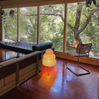 Small Clérachic tiered paper lamp lit on a wooden floor by a large window, with a daybed and a cane chair nearby.