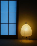 Egg-shaped Clérachic paper lamp glowing gold on the floor beside a tall window with blue panes.