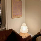 A small tiered Clérachic paper lamp lit in the corner of a room, sitting on a knitted pouffe below a framed print.