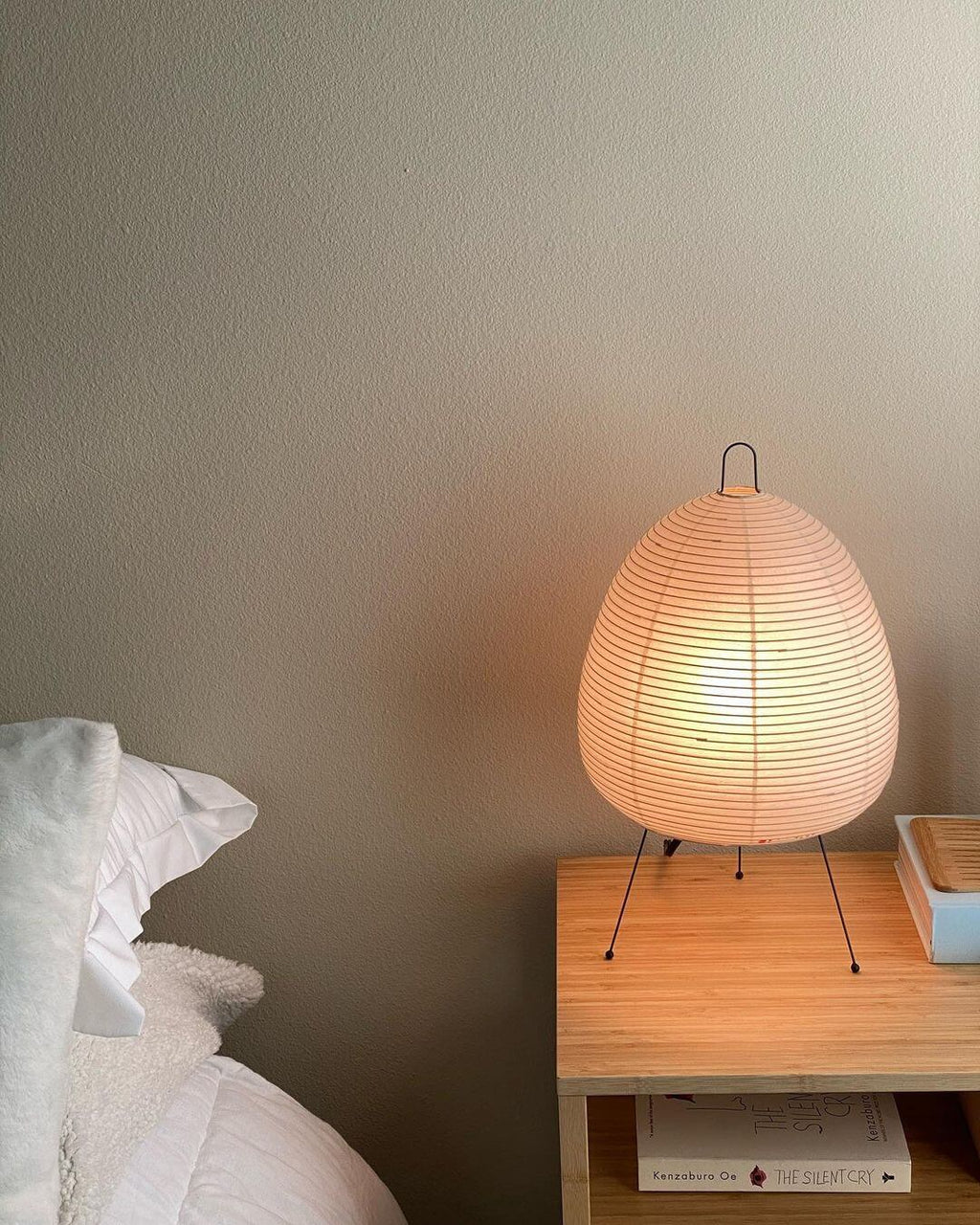 This Clérachic paper lamp glows a soft peach on a pale wooden bedside table, with white bedding at the left.