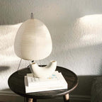 Sunlit Clérachic paper lamp on a dark round side table, with a curved ceramic vase and books beneath it.