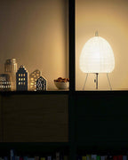 Clérachic paper lamp lit on a dark shelf beside small ceramic houses and a bowl of nuts.