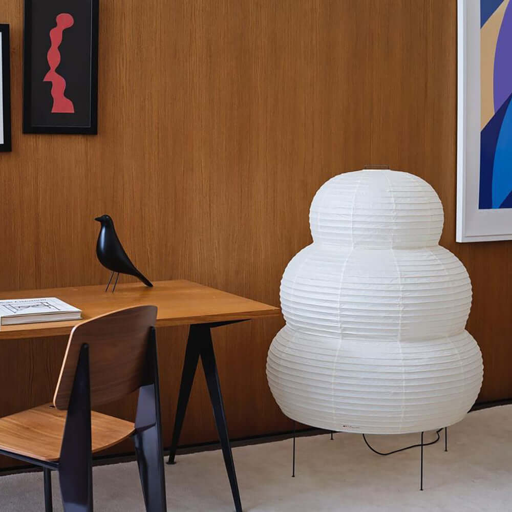 A large white tiered Clérachic paper lamp on the floor next to a wooden desk, a dark chair and a bird figurine.