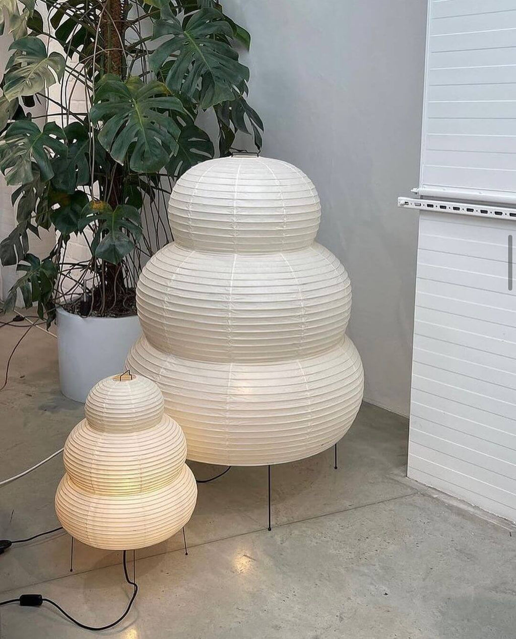 Two Clérachic tiered paper lamps, one large and one small, stand on a pale floor beside a leafy monstera plant.