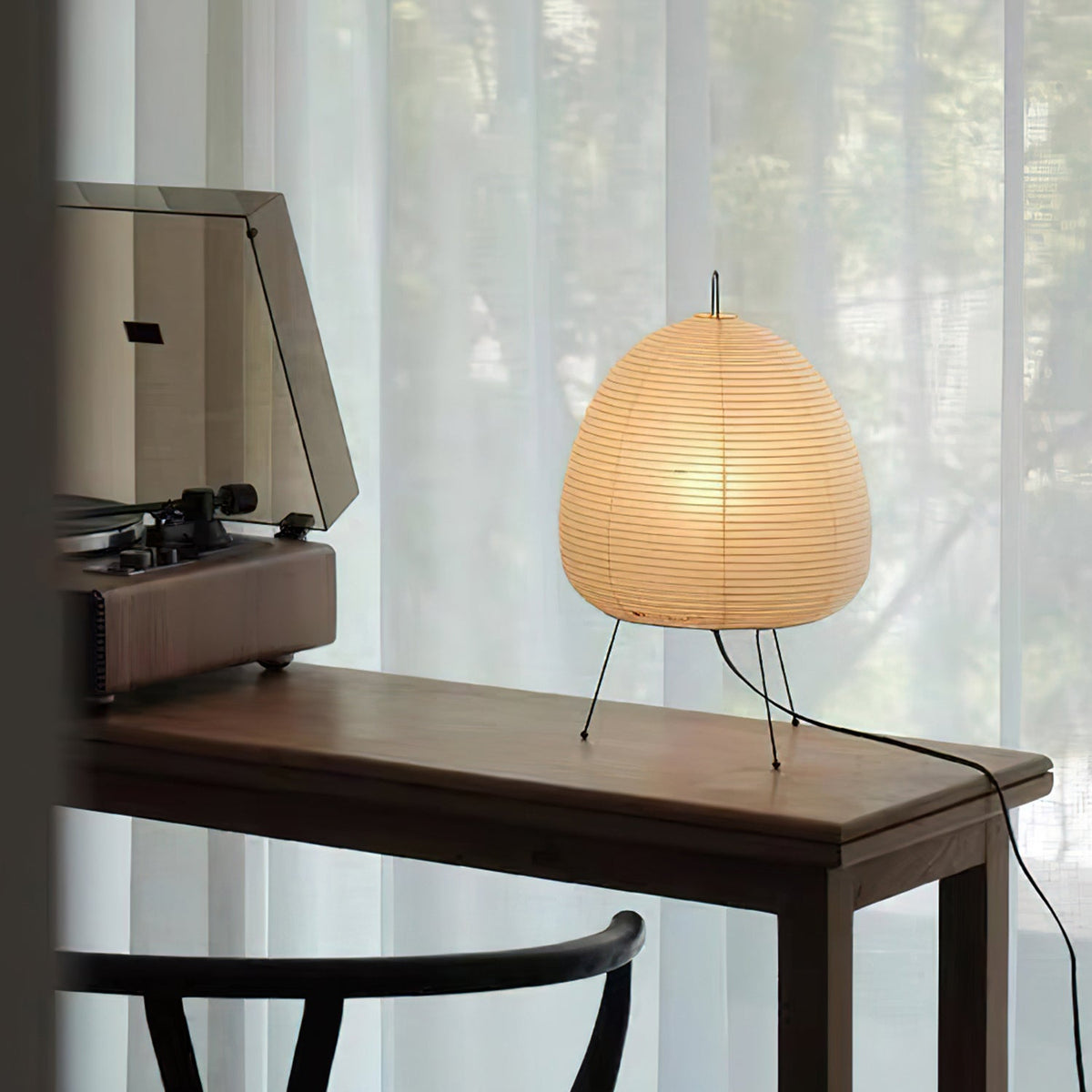 This Clérachic paper lamp lit on a dark wooden desk beside a record player, with sheer curtains behind and a black cord at the edge.