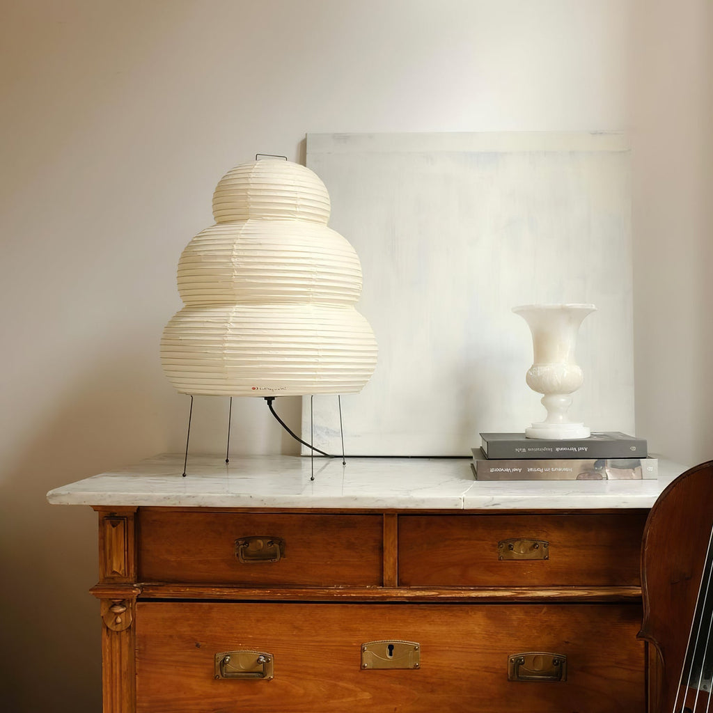 A tiered Clérachic paper lamp on a wooden chest of drawers with a marble top, beside a white vase.