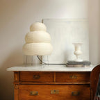 A tiered Clérachic paper lamp on a wooden chest of drawers with a marble top, beside a white vase.