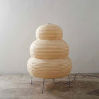 Tiered Clérachic paper lamp, unlit, on four black legs on a concrete floor, its cord running off to the right.