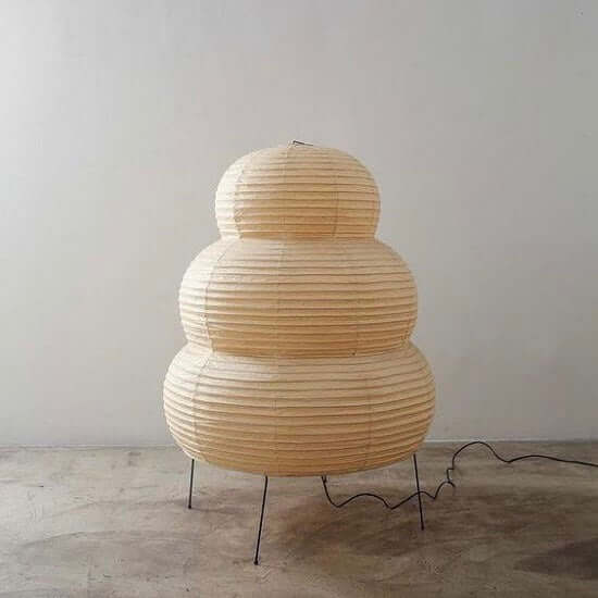 Tiered Clérachic paper lamp, unlit, on four black legs on a concrete floor, its cord running off to the right.