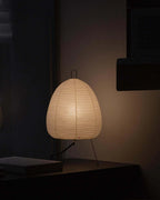 A Clérachic paper lamp glows low on a dark shelf in a dim room, with its black hanging loop visible on top.