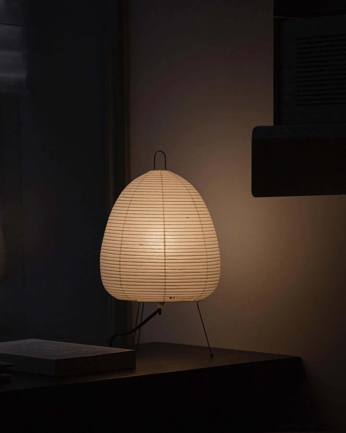 A Clérachic paper lamp glows low on a dark shelf in a dim room, with its black hanging loop visible on top.