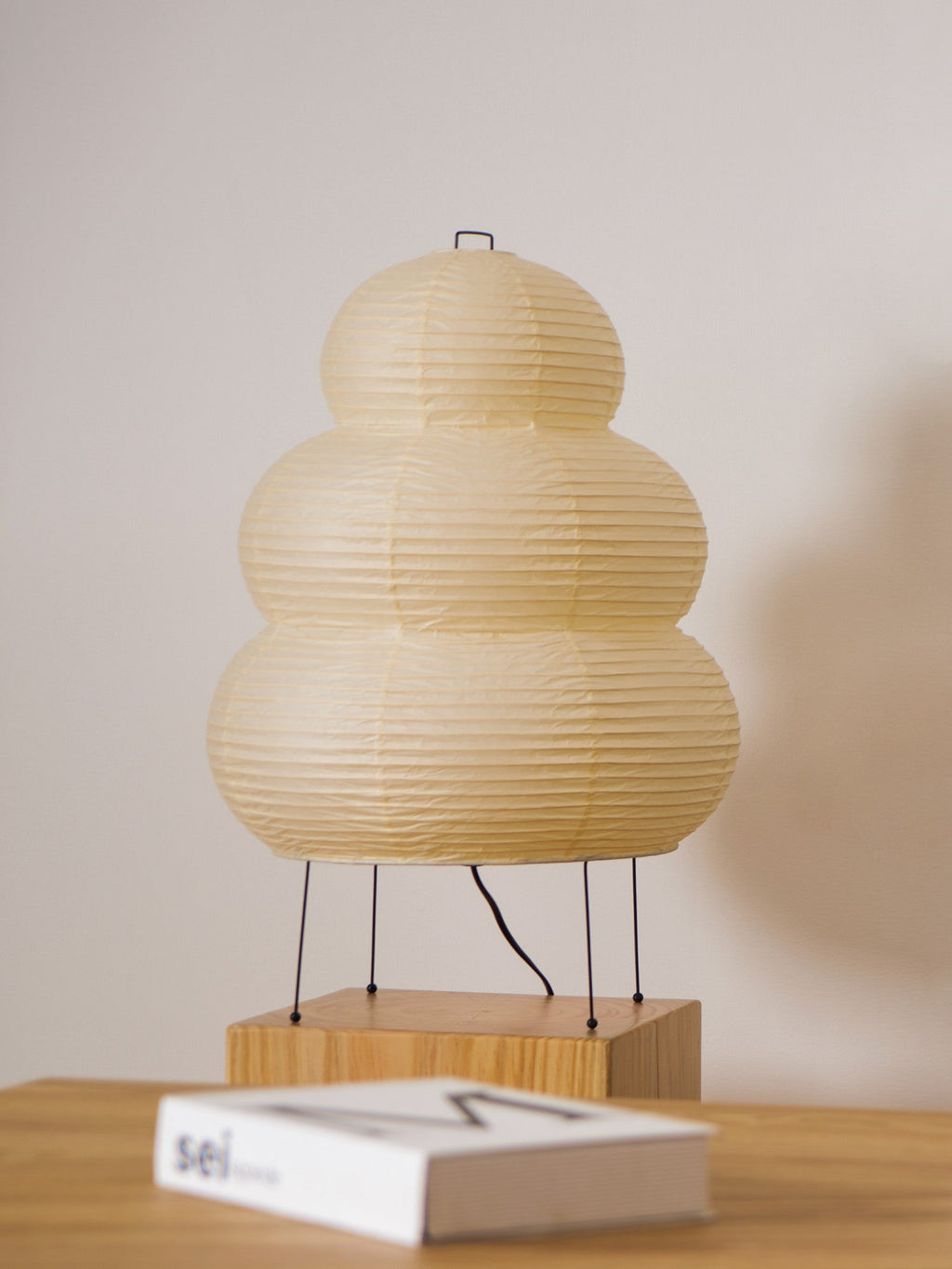 A Clérachic tiered paper lamp stands on a wooden block at a desk, with a closed white book in the foreground.