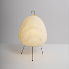 Egg-shaped Clérachic paper lamp on a plain grey backdrop, its shade finely ribbed above three black legs with round feet.
