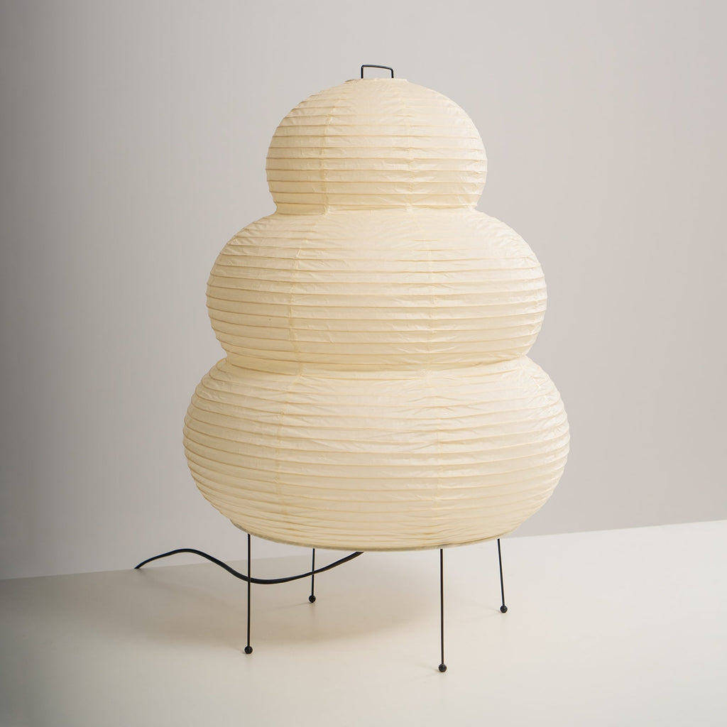 Three-quarter view of a tiered Clérachic paper lamp on four black legs, its cord curling out across a white surface.