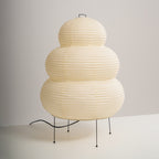 Three-quarter view of a tiered Clérachic paper lamp on four black legs, its cord curling out across a white surface.
