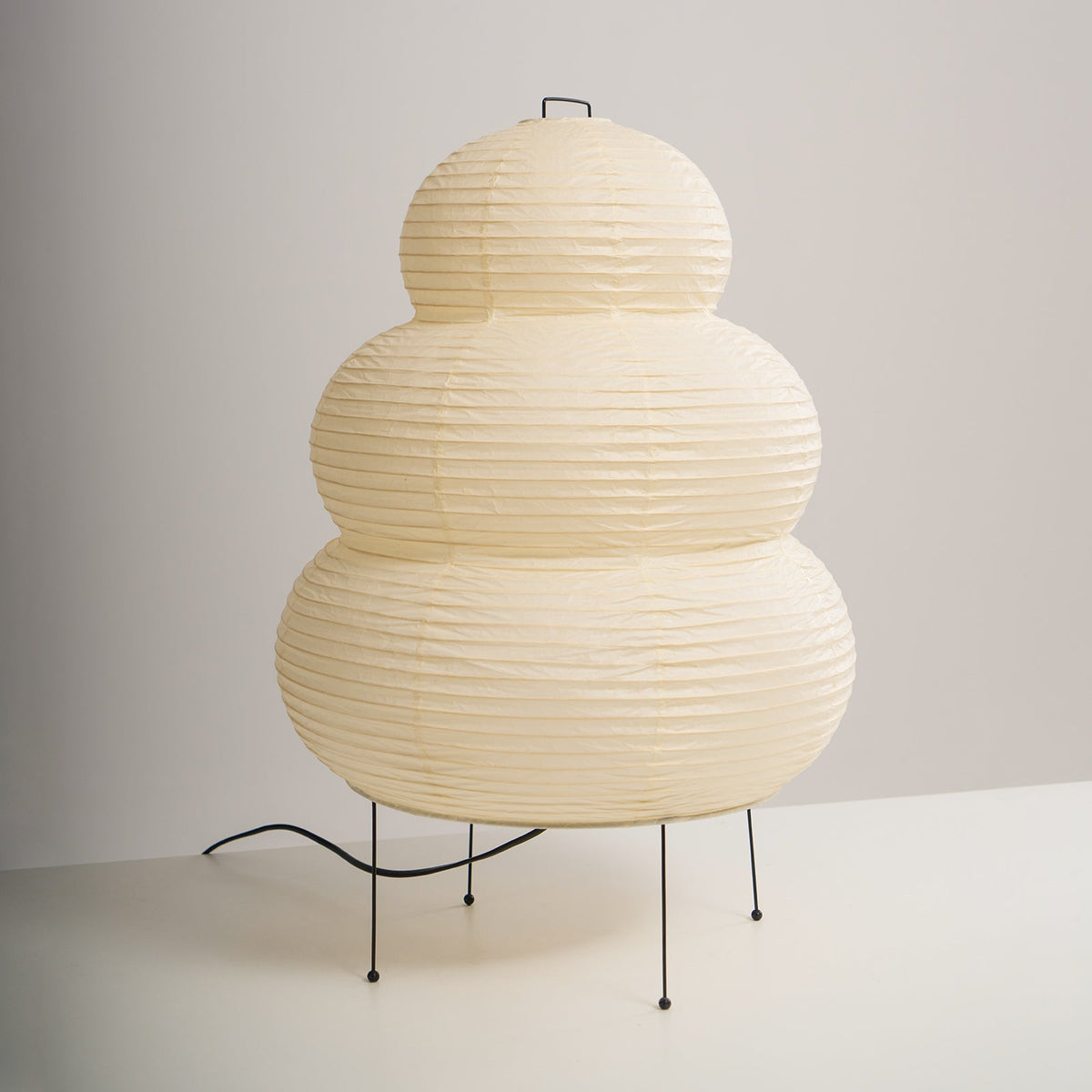 Three-quarter view of a tiered Clérachic paper lamp on four black legs, its cord curling out across a white surface.