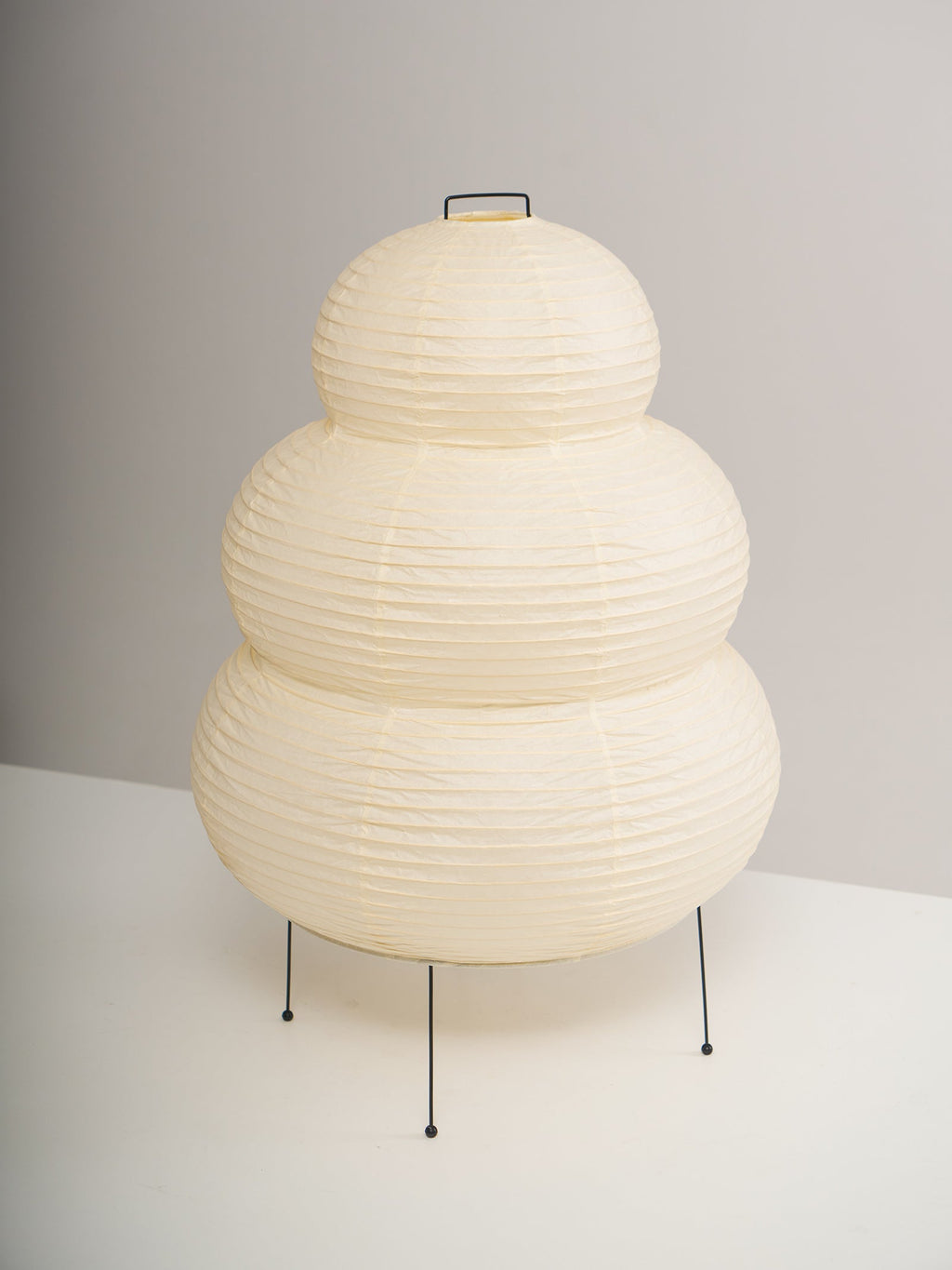 Side view of a tiered Clérachic paper lamp, switched off, on thin black legs against a plain grey wall.