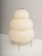 Side view of a tiered Clérachic paper lamp, switched off, on thin black legs against a plain grey wall.