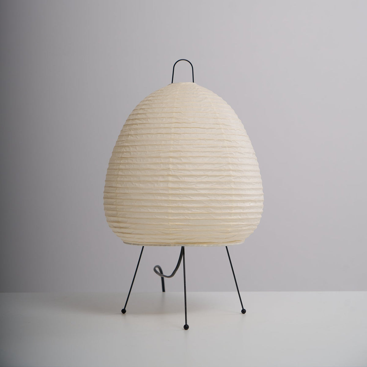 Back view of an egg-shaped Clérachic paper lamp, unlit, on three black legs with its black cord looped at the base.