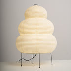 A lit Clérachic tiered paper lamp on four black legs, its cord looping across a white surface against a grey wall.