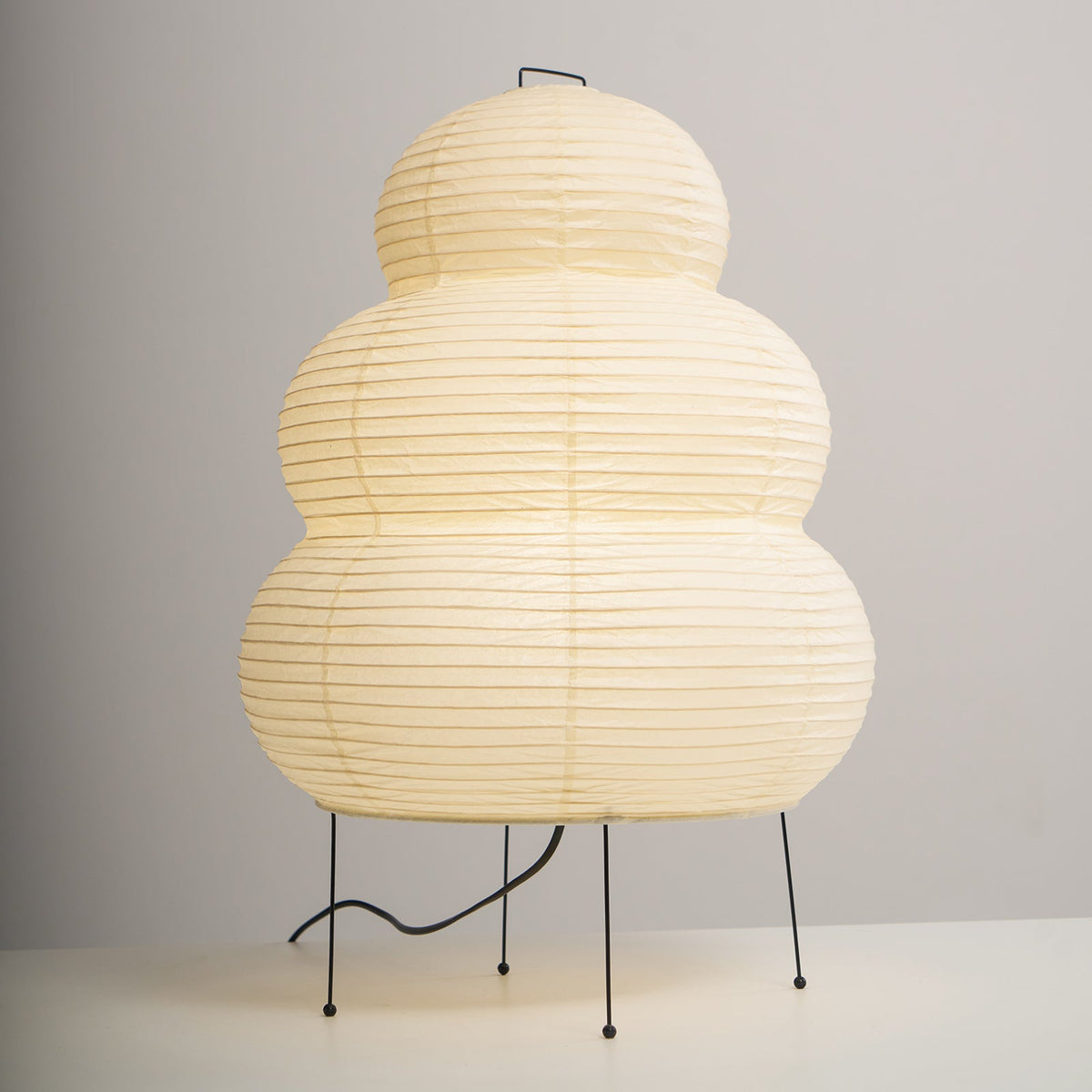 A lit Clérachic tiered paper lamp on four black legs, its cord looping across a white surface against a grey wall.