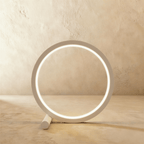 Clérachic white ring-shaped touch lamp glowing warm on its short cylindrical foot, set against a limewash wall.