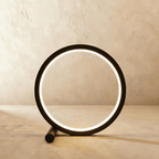 Clérachic black ring table lamp lit with a bright inner glow, standing on a short cylindrical foot against a limewash wall.