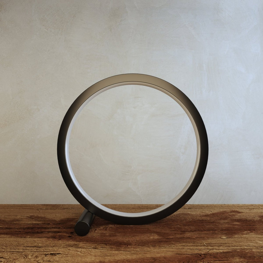 Clérachic black ring lamp switched off on a rustic wooden tabletop, with its round foot and white inner diffuser against a grey wall.
