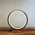 Clérachic black ring lamp lit warm on a rustic wooden tabletop, the glowing acrylic band running around the inside of the circle.