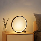 Clérachic black ring lamp glowing on a pale wood bedside table beside a glass jar of eucalyptus and a grey pillow.