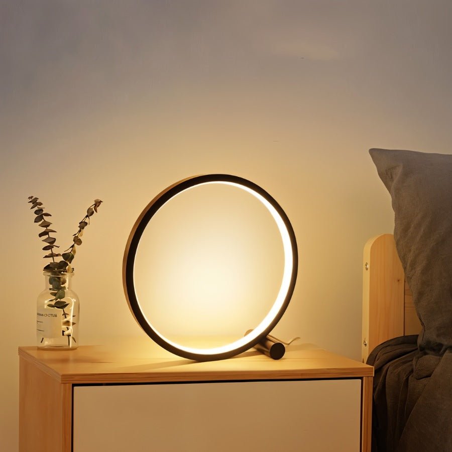 Clérachic black ring lamp glowing on a pale wood bedside table beside a glass jar of eucalyptus and a grey pillow.