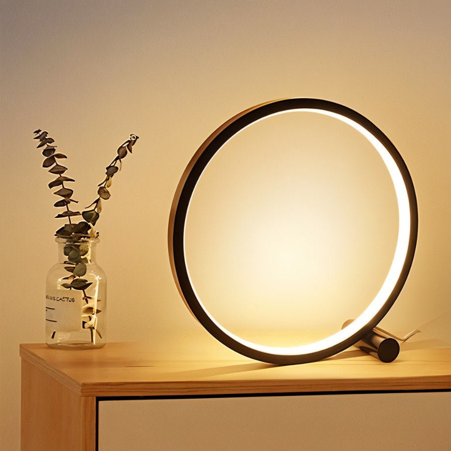 Close view of the Clérachic black ring lamp lighting a wooden bedside top, next to a glass jar of dried eucalyptus stems.