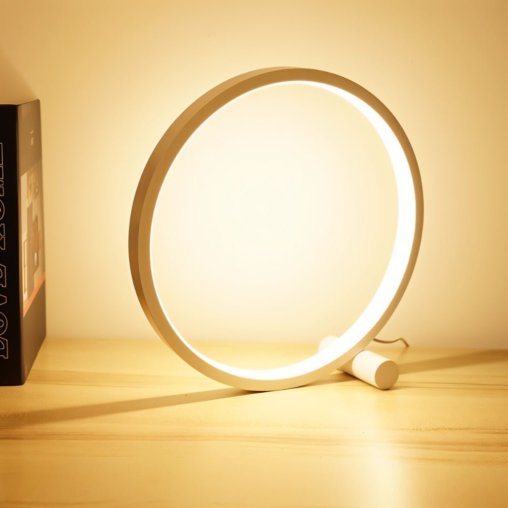 Clérachic white ring touch lamp glowing warm on a pale wooden desk beside a dark hardback book, its USB lead trailing behind.