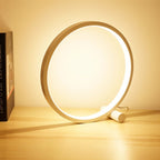 Clérachic white ring touch lamp glowing warm on a pale wooden desk beside a dark hardback book, its USB lead trailing behind.