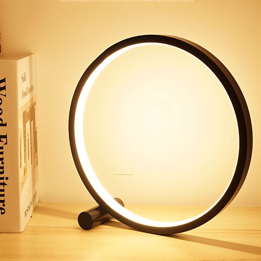 Clérachic black ring lamp lighting a wooden shelf in warm tones, beside a book on wood furniture, with a short foot at its base.