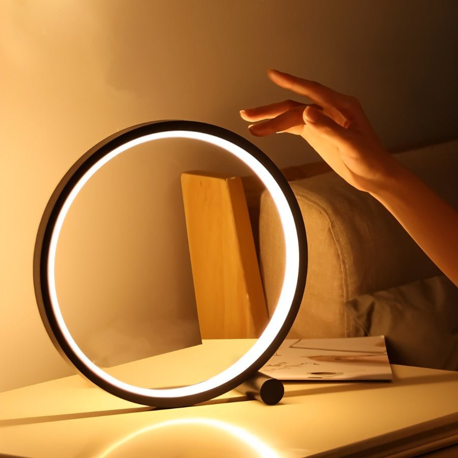 A hand reaches toward the Clérachic black ring lamp on a bedside table, showing the touch control with a cream sofa behind.