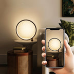 Clérachic black ring lamp glowing on a white pedestal table above a cane-sided drum, while a phone in hand photographs it.