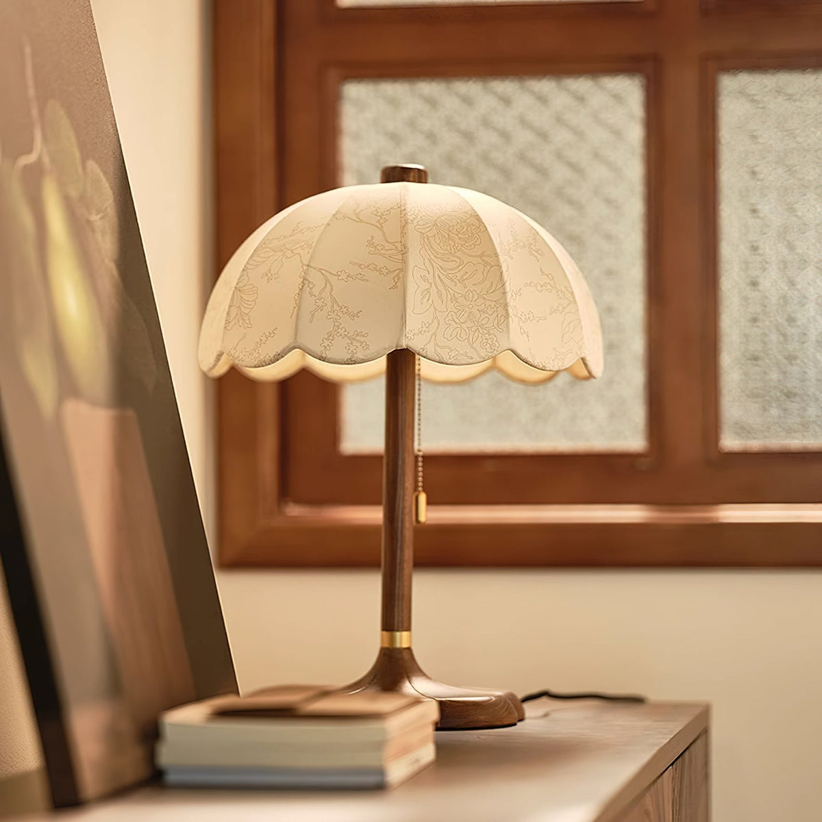 Scalloped beige shade with a faint printed pattern on this Clérachic table lamp, standing on a sideboard in front of a wood-framed window.