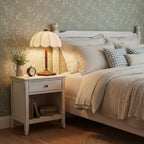 Lit bedside Clérachic table lamp on a white nightstand, with a gingham cushion and a small posy in a jug beside a made bed.