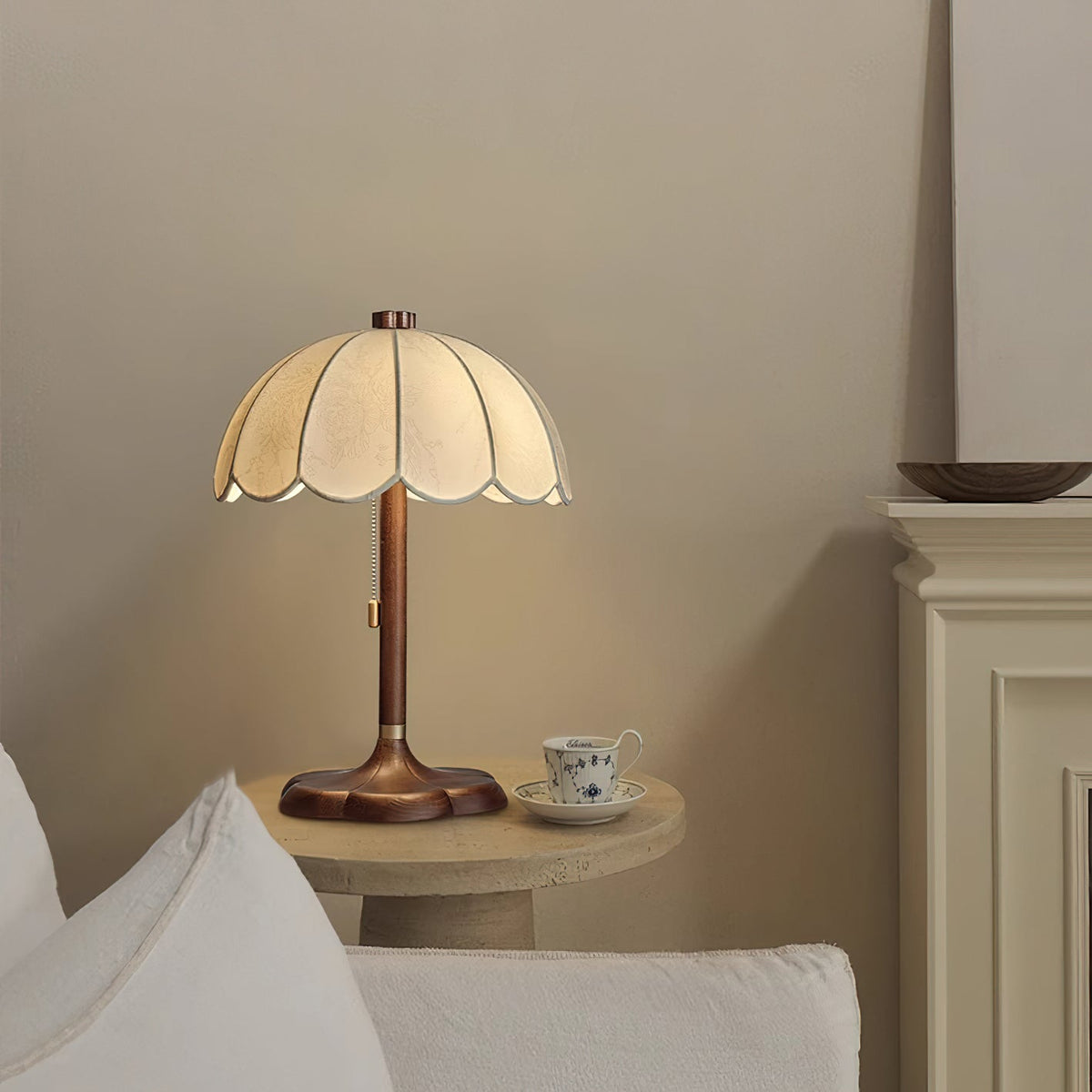 Clérachic table lamp with a scalloped shade beside a small stone table with a patterned teacup, next to a white sofa.