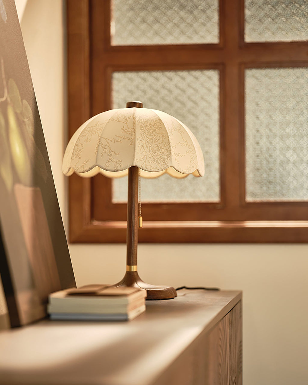 Clérachic table lamp on a wooden cabinet by a window with a patterned glass pane, with a short stack of books at its foot.