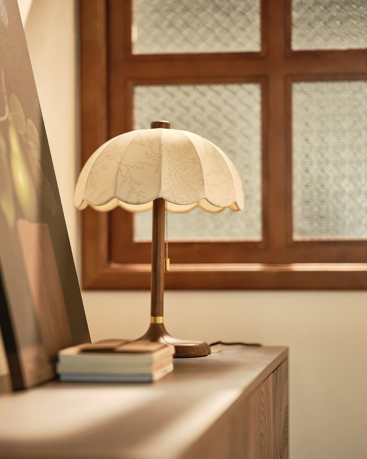 Clérachic table lamp on a wooden cabinet by a window with a patterned glass pane, with a short stack of books at its foot.
