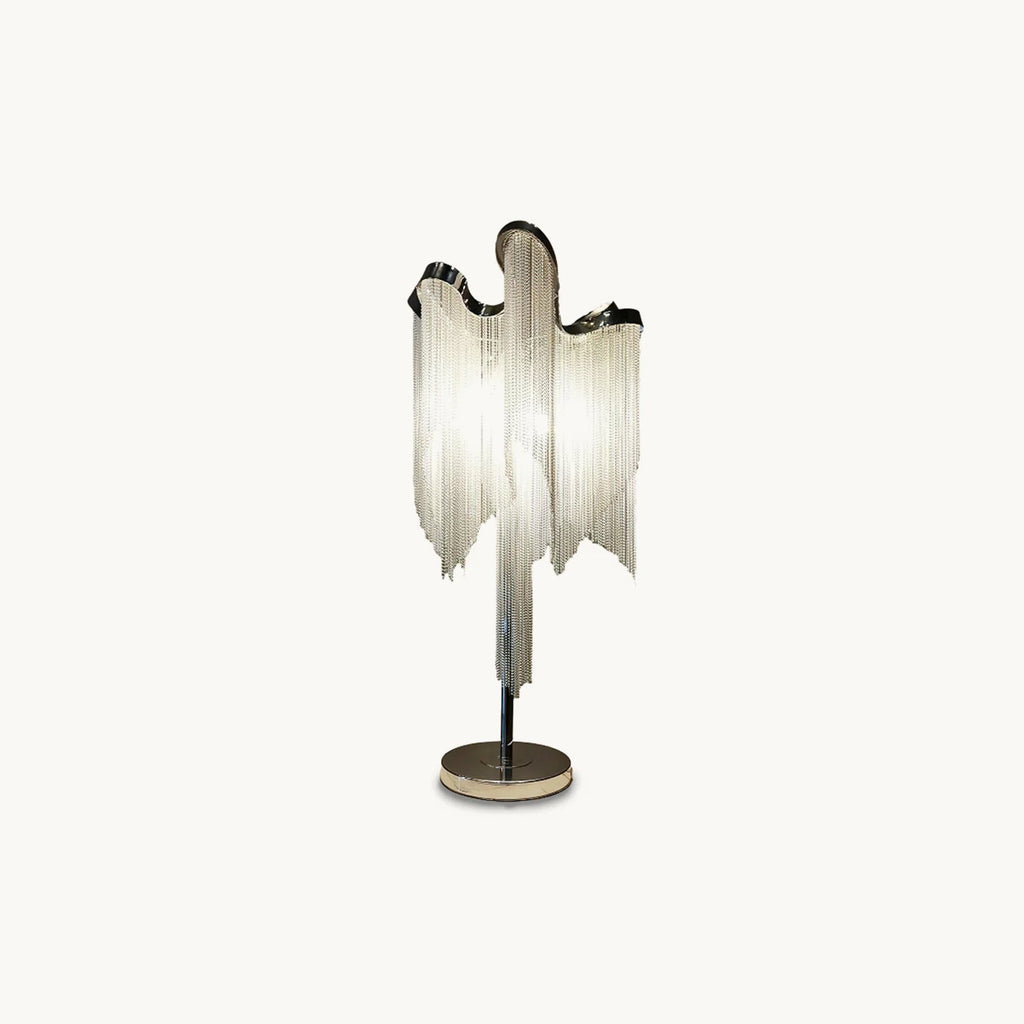 A Clérachic table lamp in silver on a plain white backdrop, its draped chain fringe falling from a curved top to a round base.