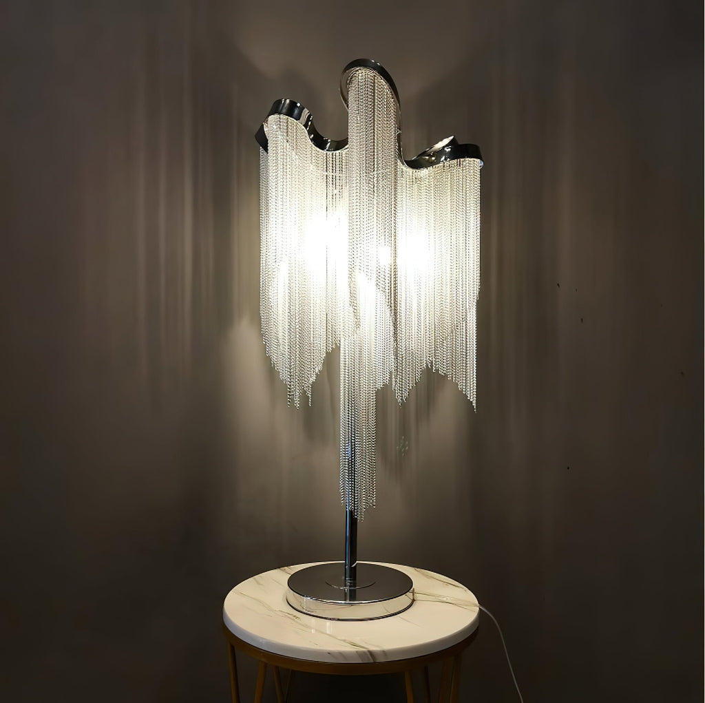 A lit Clérachic chain-fringe lamp on a round white marble-topped stand against a dark wall, its fringe glowing warm.